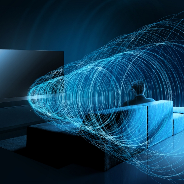 Person sitzt auf einem Sofa vor einem Fernseher, umgeben von visualisierten Klangwellen in Blau – Darstellung von Surround-Sound. Person sitzt auf einem Sofa vor einem Fernseher, umgeben von visualisierten Klangwellen in Blau – Darstellung von Surround-Sound.